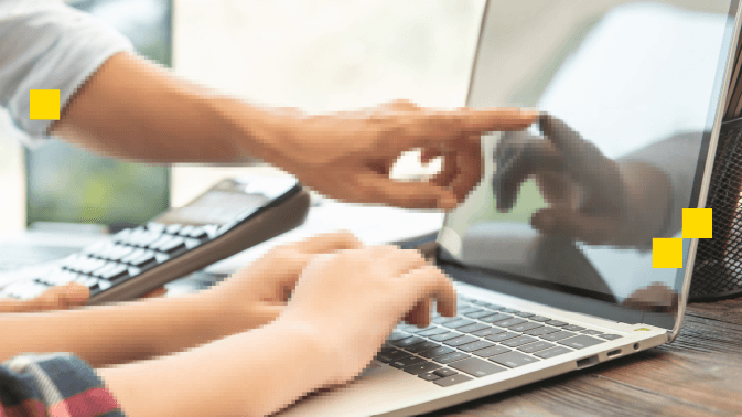 Primer plano de dos personas colaborando frente a una laptop. Una persona apunta con el dedo a la pantalla mientras la otra teclea en el teclado. En el fondo, se ve parcialmente una calculadora y un escritorio de madera, sugiriendo un entorno de trabajo o estudio. La escena transmite cooperación, enseñanza o resolución de tareas en equipo, con un enfoque en la interacción digital.